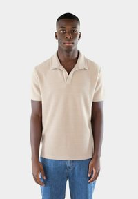 Beige kortärmad polo med en strukturerad stickad mönster, utrustad med en knäppkrage och en avslappnad passform, i kombination med blå denimjeans.