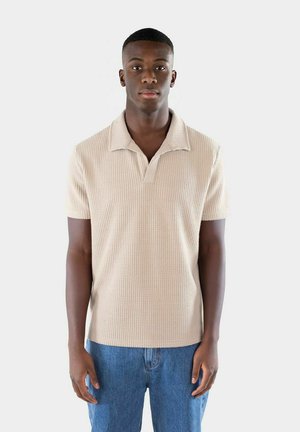Beige kortärmad polo med en strukturerad stickad mönster, utrustad med en knäppkrage och en avslappnad passform, i kombination med blå denimjeans.