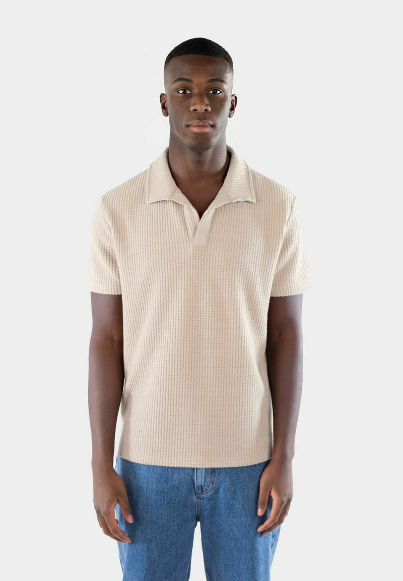 Beige kortärmad polo med en strukturerad stickad mönster, utrustad med en knäppkrage och en avslappnad passform, i kombination med blå denimjeans.