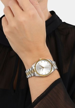 Silber- und goldfarbener Armbanduhr mit einem runden silbernen Zifferblatt, Kristallmarkierungen und einem zweifarbigen Gliederarmband, getragen an einem nackten Handgelenk.