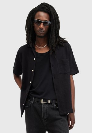Czarna koszula z guzikami, o teksturowanej tkaninie, z krótkimi rękawami i kieszenią na piersi, zestawiona z czarnym t-shirtem i ciemnymi dżinsami.
