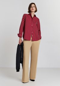 Camicia a righe rosse e bianche con maniche lunghe, abbinata a pantaloni beige a gamba larga. Giacca di pelle nera appoggiata sul braccio.