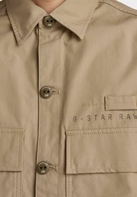 Camicia button-up beige con colletto classico, dotata di due tasche sul petto e dettagli loghi stampati. Realizzata in tessuto liscio con una finitura pulita.