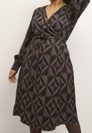 Kvinde iført en langærmet, knælængde slå-om-kjole med geometrisk sort og brun mønster, stående foran en ensfarvet baggrund.