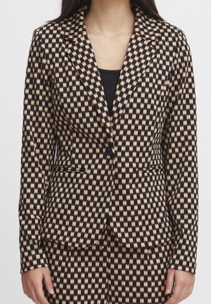 Frau trägt einen enganliegenden Blazer und Hosen mit einem beige-schwarzen Karomuster über einem schwarzen Oberteil.