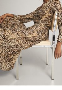 Długa sukienka w brązowo-beżowe wzory z długimi rękawami, z dekoltem w kształcie litery V i swobodnym krojem, noszona podczas siedzenia na srebrnym krześle.