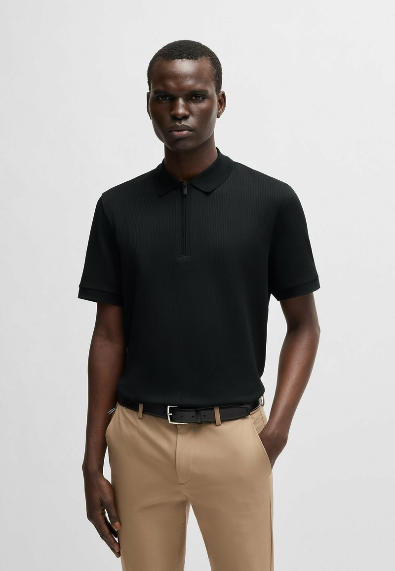 Black ribbed polo shirt with short sleeves and a zippered collar, paired with beige pants and a black belt featuring a silver buckle.