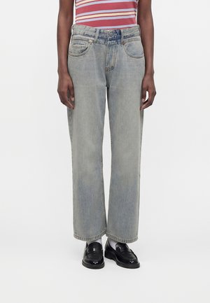 Person iført falmede grå jeans med lige ben, sorte loafers med hvide sokker og en rød- og hvidstribet skjorte, stående mod en ensfarvet baggrund.