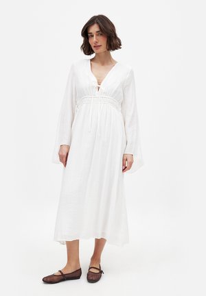 Robe longue blanche en tissu texturé, décolleté en V profond, taille à cordon, et manches évasées. Associée à des chaussures plates brun foncé.