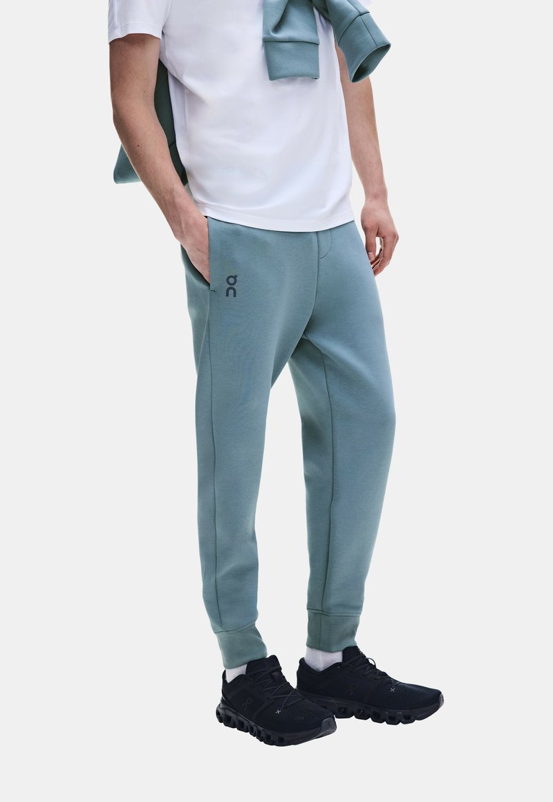Personne portant un pantalon de jogging bleu clair, des chaussettes blanches, des baskets noires et un t-shirt blanc avec un sweat bleu clair assorti drapé sur les épaules.