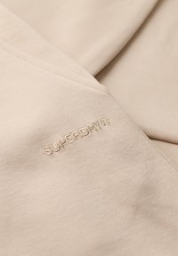 Sudadera de algodón beige con una textura suave, que presenta un logo bordado de "SUPERDRY" en un beige ligeramente más oscuro. Diseño simple y minimalista.