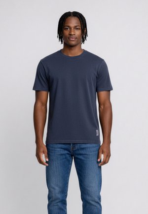 Giovane con capelli intrecciati che indossa una t-shirt blu scuro semplice e jeans blu, in piedi davanti a uno sfondo grigio chiaro.