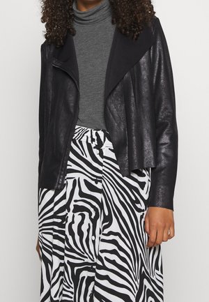 Personne aux cheveux bouclés portant un col roulé gris, une veste en cuir noire et une jupe à rayures zèbre noire et blanche avec des boutons.