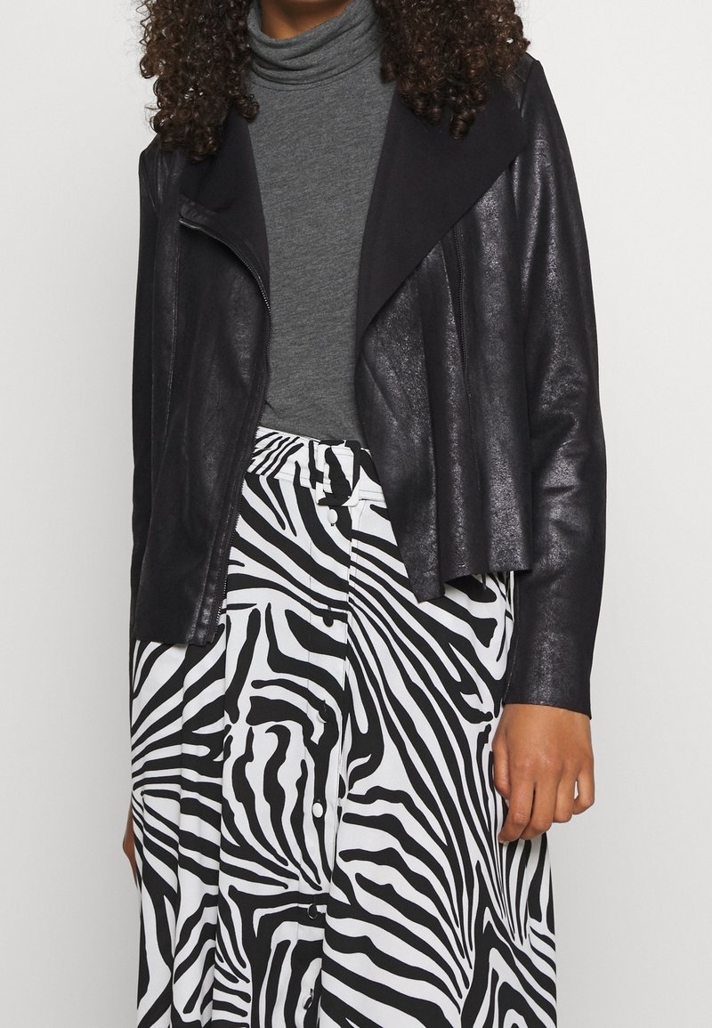 Personne aux cheveux bouclés portant un col roulé gris, une veste en cuir noire et une jupe à rayures zèbre noire et blanche avec des boutons.