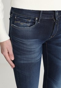 Vaqueros de mezclilla azul oscuro con corte ajustado, que presentan un ligero desgaste, dos bolsillos frontales y costuras detalladas cerca de los bolsillos.