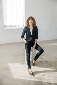 Mujer con cabello rizado que lleva un traje negro y bailarinas marrones puntiagudas, sentada sobre un bloque blanco en una habitación luminosa y minimalista con suelo de cemento.