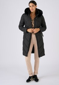 Zwarte donzen jas met een capuchon gevoerd met nepbont, in een diamantvormig doorgestikt patroon, voorzien van een ritssluiting aan de voorkant, gedragen over een luipaardprint top en beige broek.