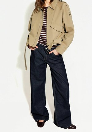 Beige cropped jack met twee voorkruiszakken, gecombineerd met een donker gestreept shirt en een wijd uitgesneden donkere jeans. Bruine flats maken de outfit compleet.