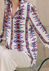 Une personne portant une chemise blanche à manches longues avec des motifs géométriques rouges, bleus et noirs, assise sur une chaise transparente avec un pantalon blanc texturé.