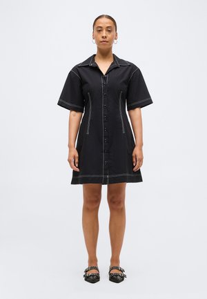 Femme debout portant une robe noire à manches courtes avec des boutons et des coutures blanches, des boucles d'oreilles en forme d'anneau et des chaussures noires à pointes ornées de clous.