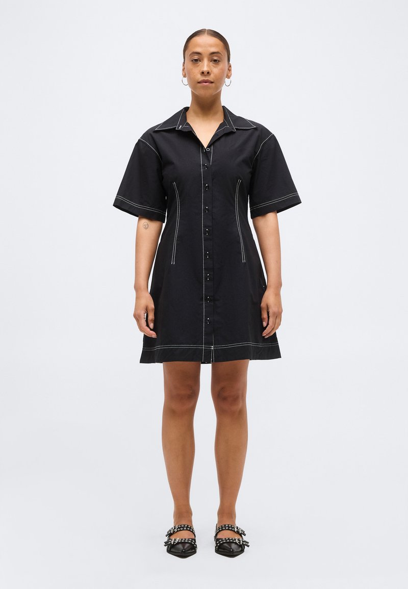 Γυναίκα όρθια που φοράει μαύρο φόρεμα με κοντά μανίκια και κουμπιά, με λευκή ραφή, σκουλαρίκια κρίκους και μαύρα μυτερά παπούτσια με νύχια.