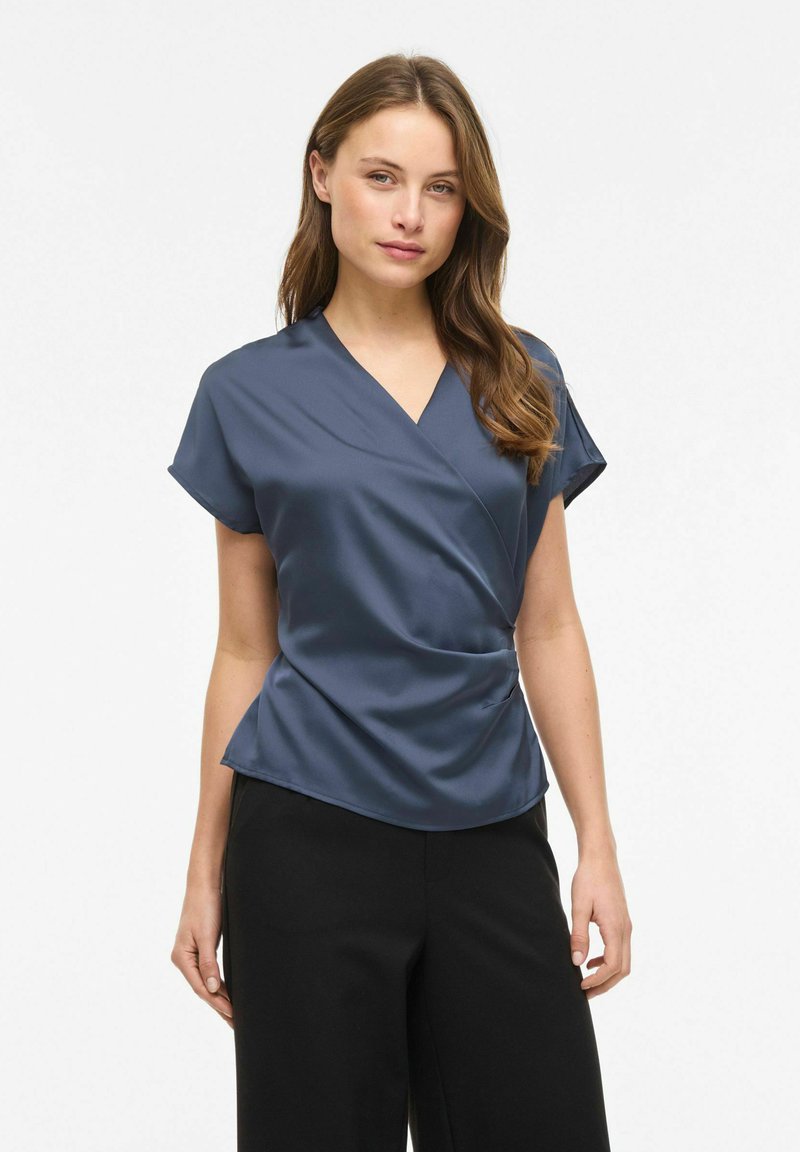 Femme à la peau claire et aux cheveux châtain clair, portant un chemisier bleu marine à manches courtes et un pantalon noir, debout devant un fond blanc uni.