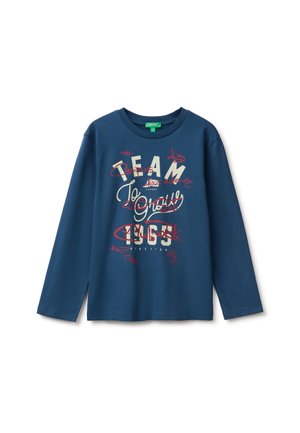 Camisa azul de manga larga con gráfico de texto en crema y rojo que dice "TEAM 1963" y otras palabras garabateadas en el frente.