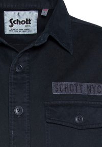 Camicia nera con bottoni e colletto, dotata di taschino sul petto, toppa in tessuto con la scritta "SCHOTT NYC" ricamata e materiale dalla consistenza liscia.