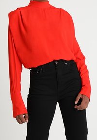 Blouse rouge à manches longues avec col montant, présentant une coupe ample et un tissu texturé, associée à un jean noir taille haute.