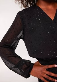 Černá průhledná blůza s dlouhými rukávy, která má texturovaný povrch s malými, lesklými tečkami a nařasenými manžetami. Výstřih do V přidává na detailu.
