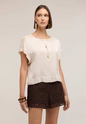 Nainen, joka pukeutunut kermaan väriseen lyhythihainen puseroon, tummanruskeisiin pitsishortseihin, kultaisiin renkaan muotoisiin korvakoruihin, kerrokselliseen kaulakoruun ja puisiiin rannekoruihin, seisoo neutraalia taustaa vasten.