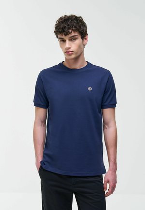 Joven con cabello rizado que lleva una camiseta azul marino con un pequeño logo circular en el pecho, de pie contra un fondo claro y uniforme.