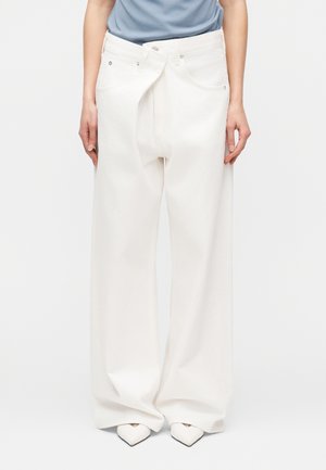 Széles szárú, fehér nadrágot viselő személy, mely elöl hasítékkal és átfedéssel rendelkezik, valamint hegyes orrú fehér cipőt visel, egyszínű háttér előtt áll.