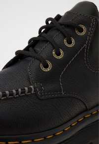Zapato de cuero negro con cordones redondos negros, ojales dorados, costuras grises y una suela gruesa negra con un acento amarillo. Detalles de superficie texturizada.