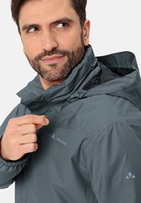 Giacca impermeabile grigio scuro con colletto alto, cappuccio regolabile e polsini elastici. Presenta un logo sul petto e sulla spalla.