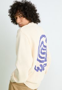 Personne avec des cheveux bouclés portant un pull en tricot crème avec un grand graphique abstrait violet dans le dos, associé à un pantalon marron clair.