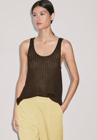 Bruine gebreide tanktop met open steekpatroon, ronde halslijn en verlengde zijkanten, gecombineerd met lichtgele linnen broek.