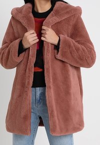 Person klädd i en dammig rosa plyschfaux fur-kappa över en svart och röd mönstrad tröja och ljusblå jeans, som håller i kragen på kappan.