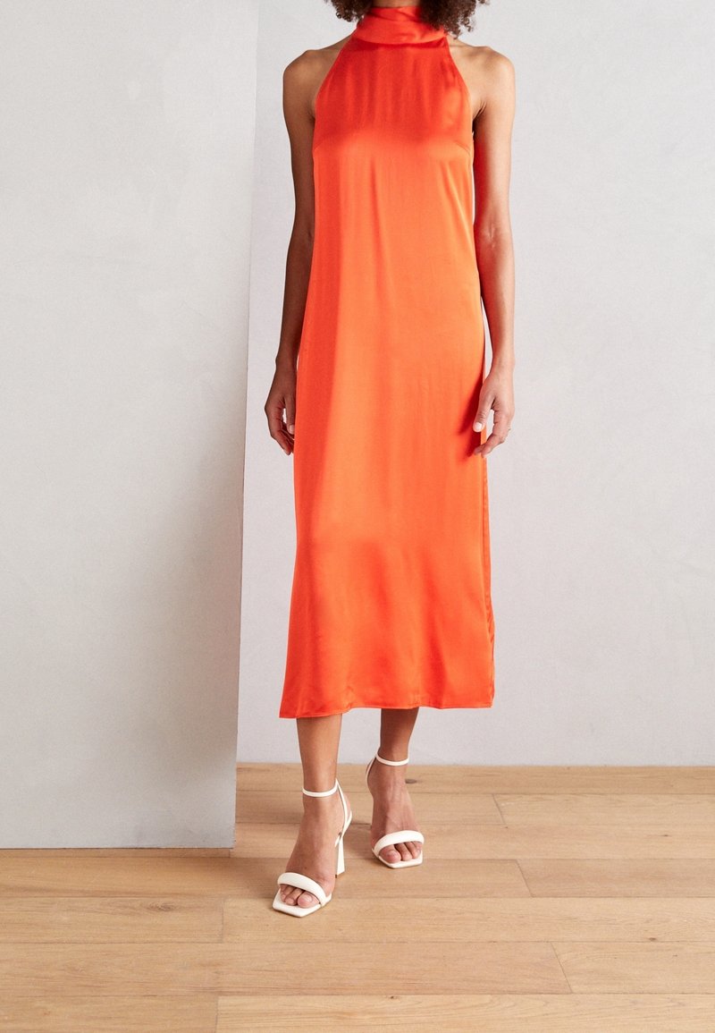 Frau in einem ärmellosen, leuchtend orangefarbenen Midikleid mit Halterneck, kombiniert mit weißen High Heels, steht auf einem Holzfußboden.