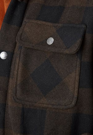 Gros plan sur un tissu à carreaux marron et noir avec une poche à rabat dotée d'un bouton-pression argenté portant l'inscription "Von Dutch California".