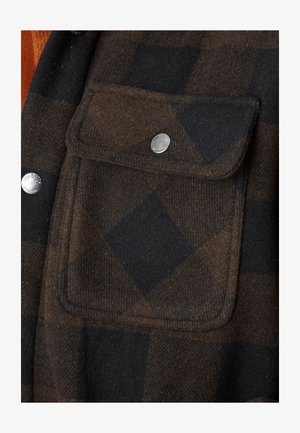 Gros plan sur un tissu à carreaux marron et noir avec une poche à rabat dotée d'un bouton-pression argenté portant l'inscription "Von Dutch California".