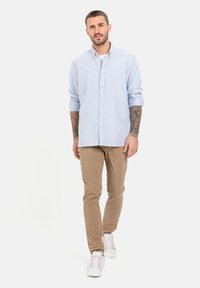 Lichtblauw gestreept overhemd met lange mouwen, borstzak, beige chino's en witte sneakers, staande op een effen achtergrond.