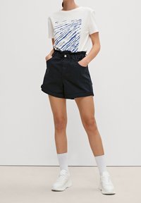 T-shirt blanc avec un motif de lignes bleues, associé à un short marine avec une taille élastique et des revers, porté avec des sneakers blanches et des chaussettes.