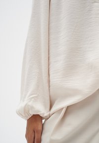 Helle beigen Bluse mit strukturiertem Finish, ausgestattet mit voluminösen Ärmeln, die an den Bündchen gerafft sind. Der glatte Stoff fällt sanft.