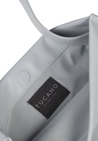 Grijze draagtas met een zachte, matte textuur; voorzien van een stoffen label met "TUCANO, MILANO 1983" in zwart. Schoon, modern interieurontwerp.