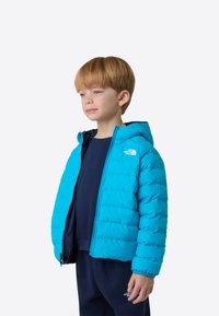 Ανοιχτό μπλε μπουφάν τύπου puffer με κουκούλα, με καπιτονέ υφή και λογότυπο. Συνδυασμένο με σκούρο πουκάμισο, σε απλό φόντο.