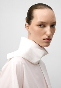 Camicia oversize rosa chiaro, con un alto colletto bianco, texture morbida, dettagli sulle cuciture laterali e design minimalista.
