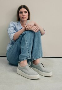 Světle šedé slip-on boty z veluru, s hrubou bílou podrážkou a jemnými švy, v kombinaci s modrými džíny.