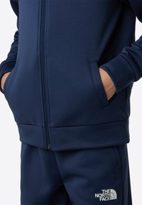 Marineblå glidelåsjakke med glatt tekstur og sidelommer. Har en logo på venstre bryst og matchende bukser.