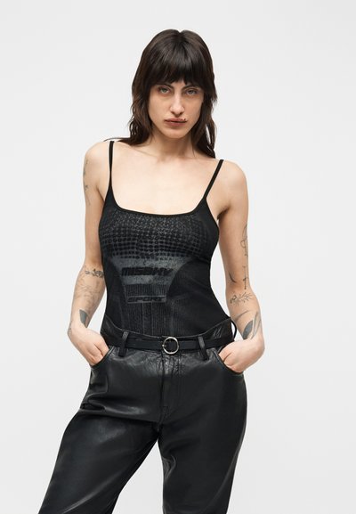 Femme aux cheveux foncés et tatouages, portant un débardeur noir à motifs et un pantalon en cuir noir, debout les mains dans les poches devant un fond blanc.